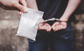 A hand holding a bag with white powder in front of a person in handcuffs
